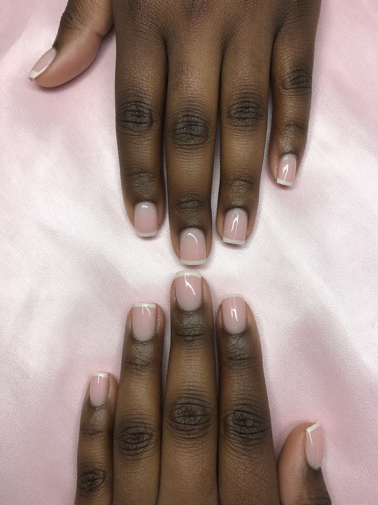 Overhead view of black hands with very short, square nails resting on a pale pink silk. The design features two vertical stripes of sheer blush polish on the sides of each nail, with a clear, natural stripe down the middle. The light is even and soft, highlighting the negative space.