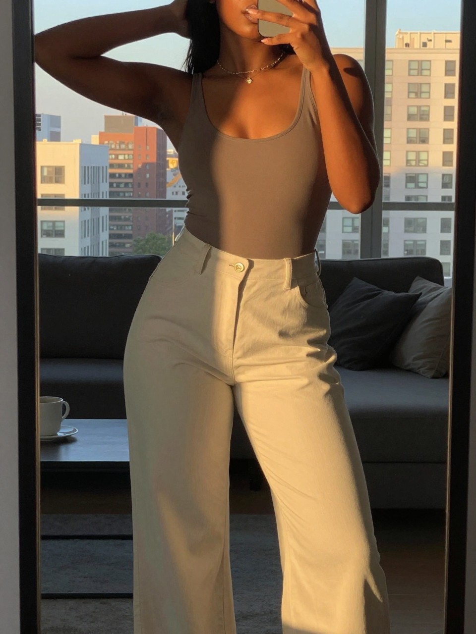 mirror selfie of a black woman in a cutout bodysuit and palazzo pants, one hand behind head, cropped at NECK so face doesn't show, in a city apartment during evening golden hour, iPhone photo quality.