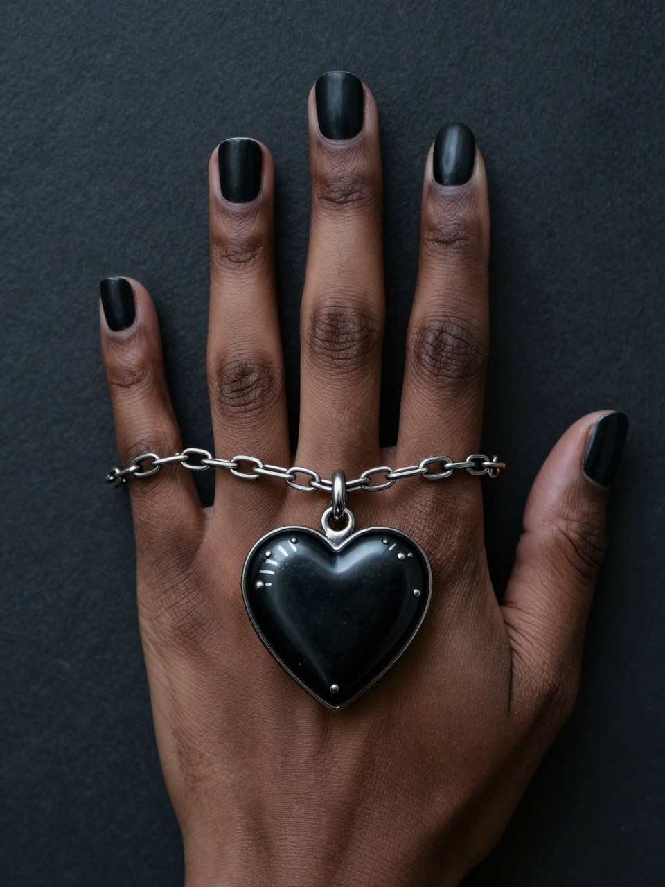 Overhead view of a black feminine hand, focusing on the ring finger. A silver chain is painted across a matte black nail, connecting to a detailed black heart locket. The other nails have simple chain links. The setting is moody and Victorian-inspired.