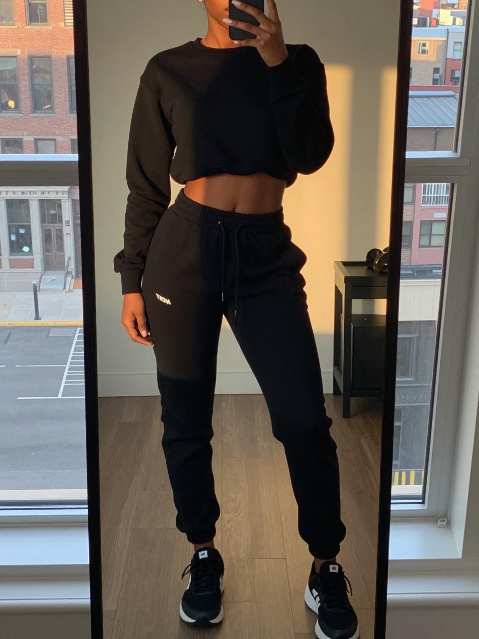 iPhone mirror selfie of a black woman in a black sweatsuit and Timbs, relaxed pose, cropped at NECK so face doesn't show, in a city apartment during evening golden hour, iPhone photo quality.
