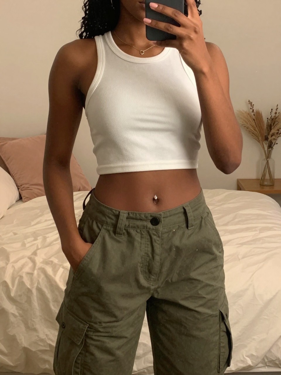 iPhone mirror selfie of a black woman in a white crop top and olive cargos with Timbs, hand in pocket, cropped at NECK so face doesn't show, in a feminine-themed bedroom with warm moody lighting, iPhone photo quality.