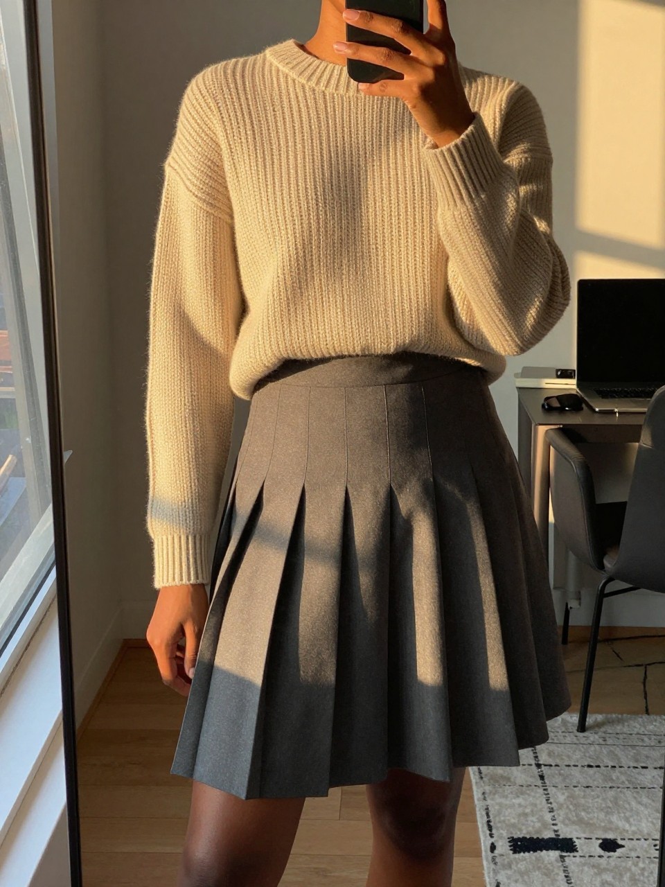 iPhone mirror selfie of a black woman in a chunky sweater and pleated skirt with Timbs, angled slightly to show the outfit proportions, cropped at NECK so face doesn't show, in a city apartment during evening golden hour, iPhone photo quality.