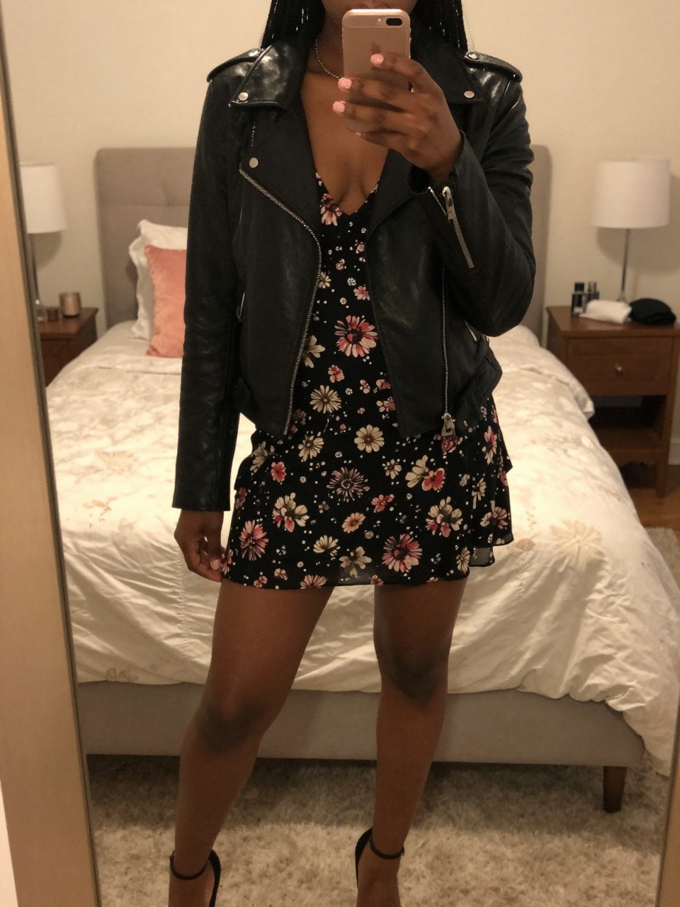 iPhone mirror selfie of a Black woman wearing a black leather jacket over a floral dress, standing with one foot forward and phone held casually, cropped at NECK so face doesn't show, in a feminine-themed bedroom, warm moody lighting, iPhone photo quality.