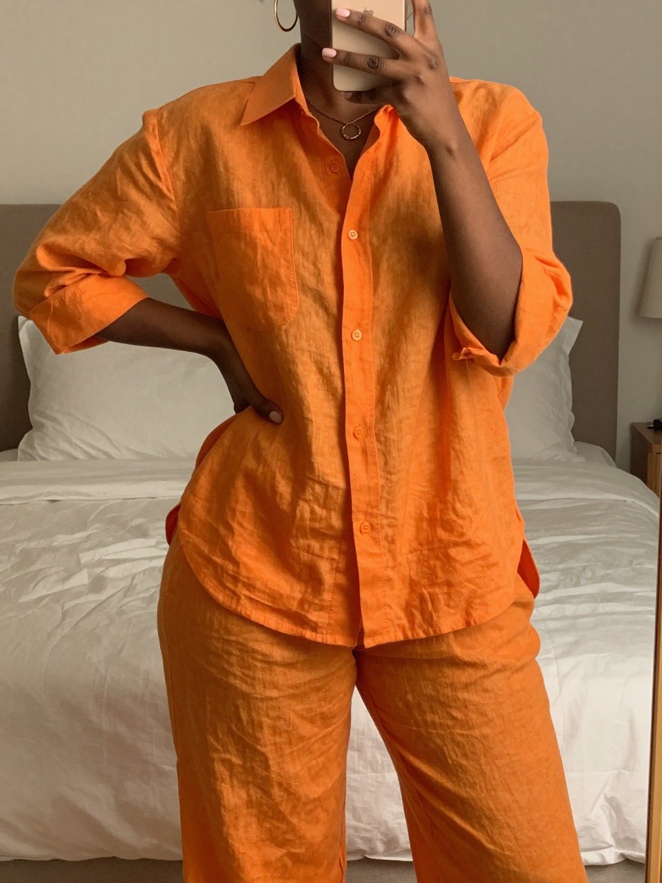iPhone mirror selfie of a black woman wearing a bright orange linen matching set with gold hoops, posing with one hand on her hip, cropped at NECK so face doesn't show, in a feminine-themed bedroom with warm moody lighting, iPhone photo quality.