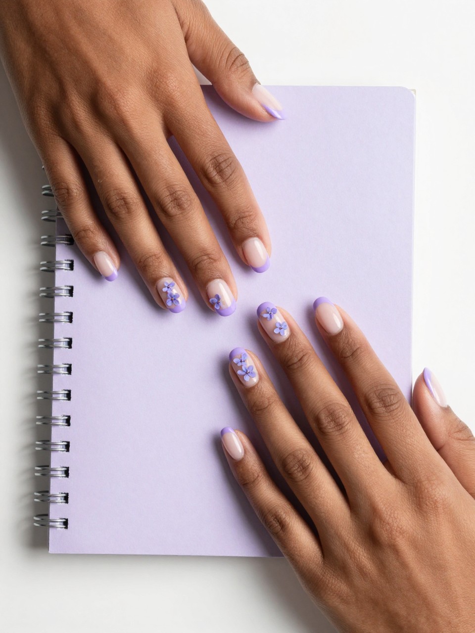 Overhead view of black feminine hands with short, rounded nails on a pastel-colored notebook. The French tips are a soft lavender. On the nude base of each nail, two or three micro lavender flowers are scattered. The overall feel is soft, modern, and coordinated.