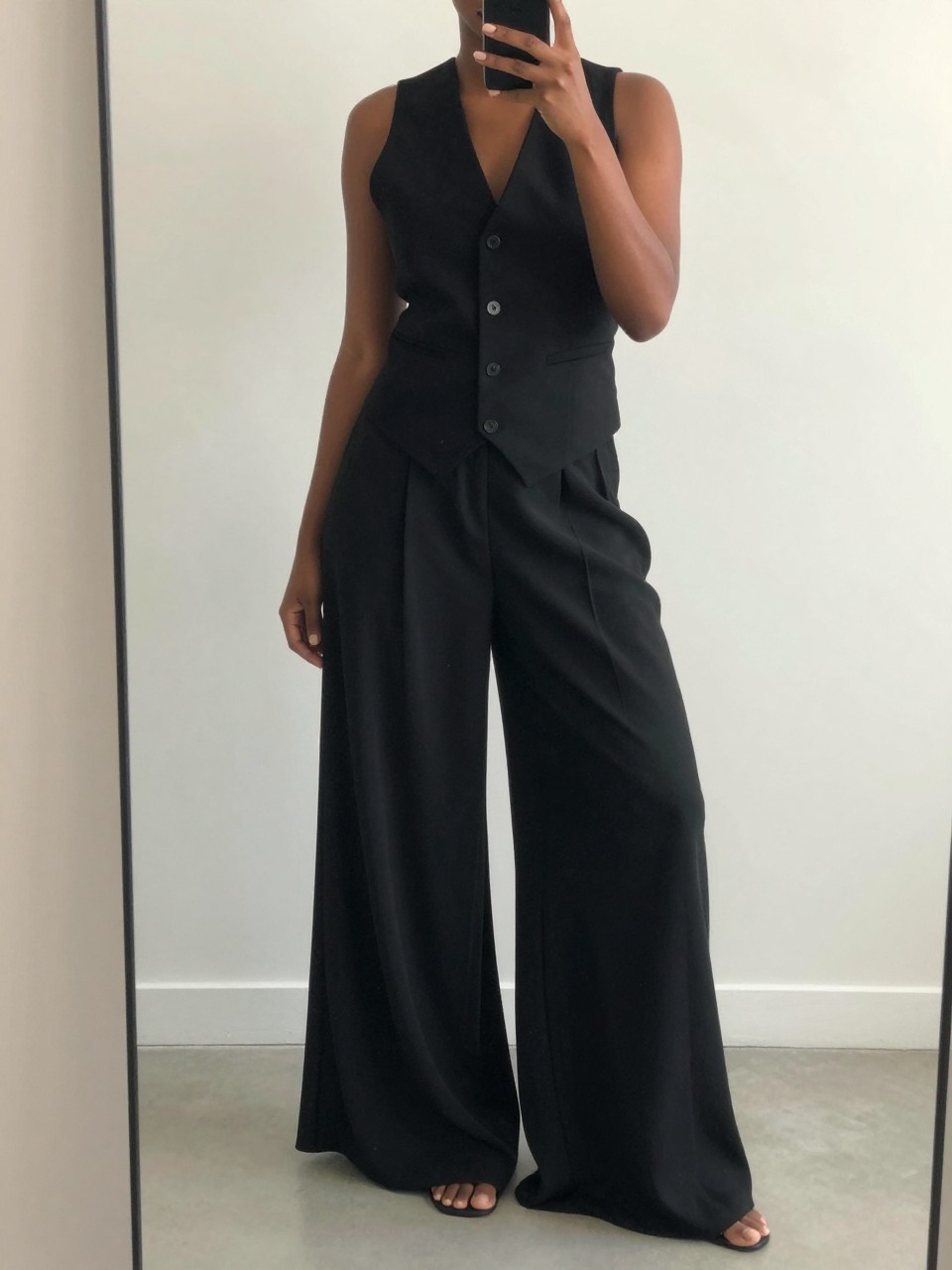 iPhone mirror selfie of a black woman wearing a black vest with flowing palazzo pants, posed with her phone low and body angled, cropped at NECK so face doesn't show, iPhone photo quality.