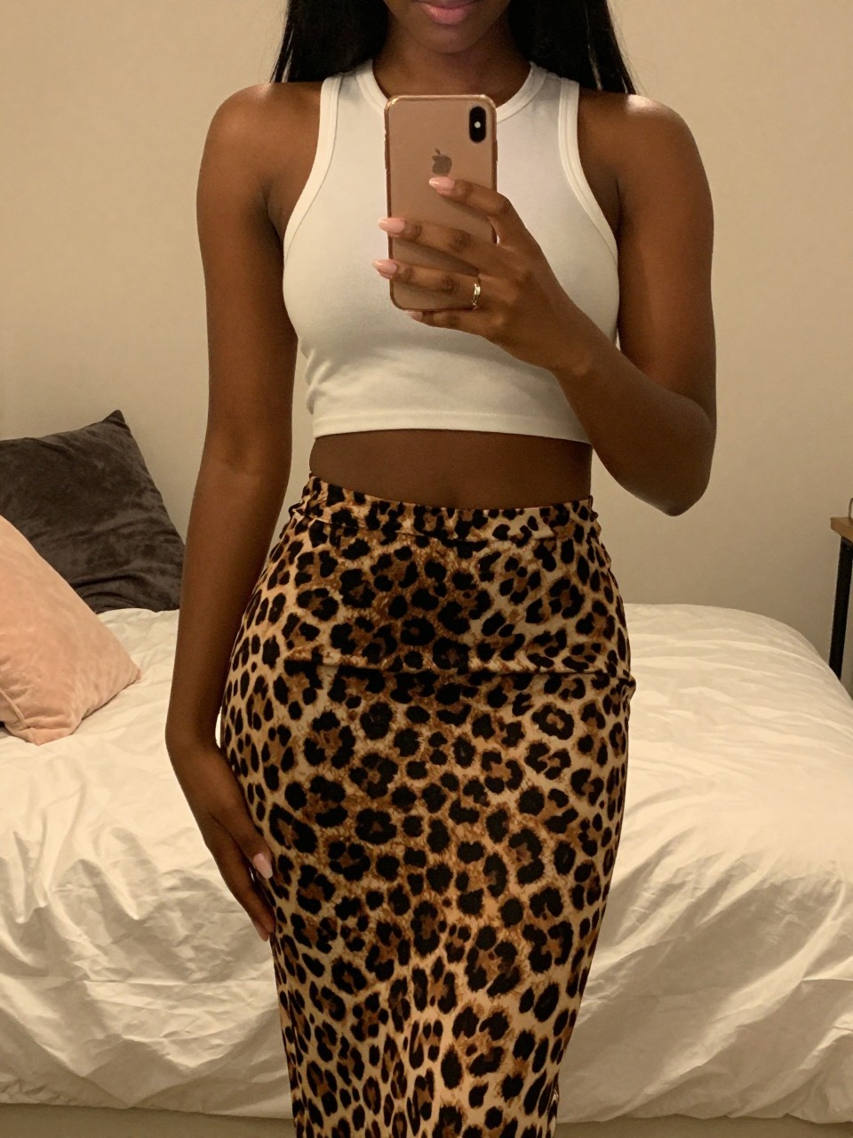 iPhone mirror selfie of a black woman in a leopard midi skirt and white crop top, holding her phone at chest level, cropped at NECK so face doesn't show, in a feminine-themed bedroom with warm moody lighting, iPhone photo quality.