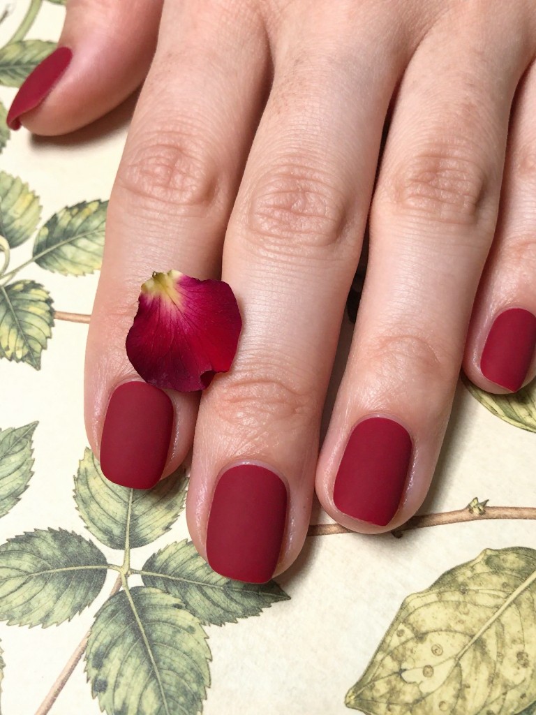 Macro, top-down view of hands over a vintage botanical print. The nails are matte crimson. The ring fingers have a real, delicate pressed red rose petal or tiny rosebud encapsulated under glossy gel. The light highlights the natural texture.