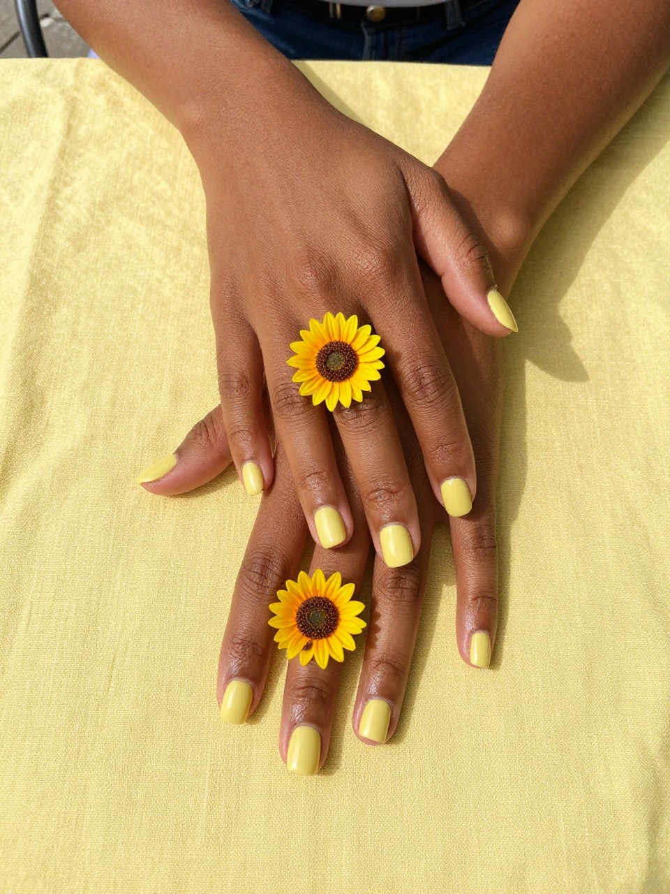 Overhead, top-down view of black feminine hands with short, rounded nails resting on a yellow linen tablecloth. The nails are a glossy butter yellow. Each ring finger features a large, detailed sunflower with a textured brown center. The setting is a sun-drenched outdoor cafe table.