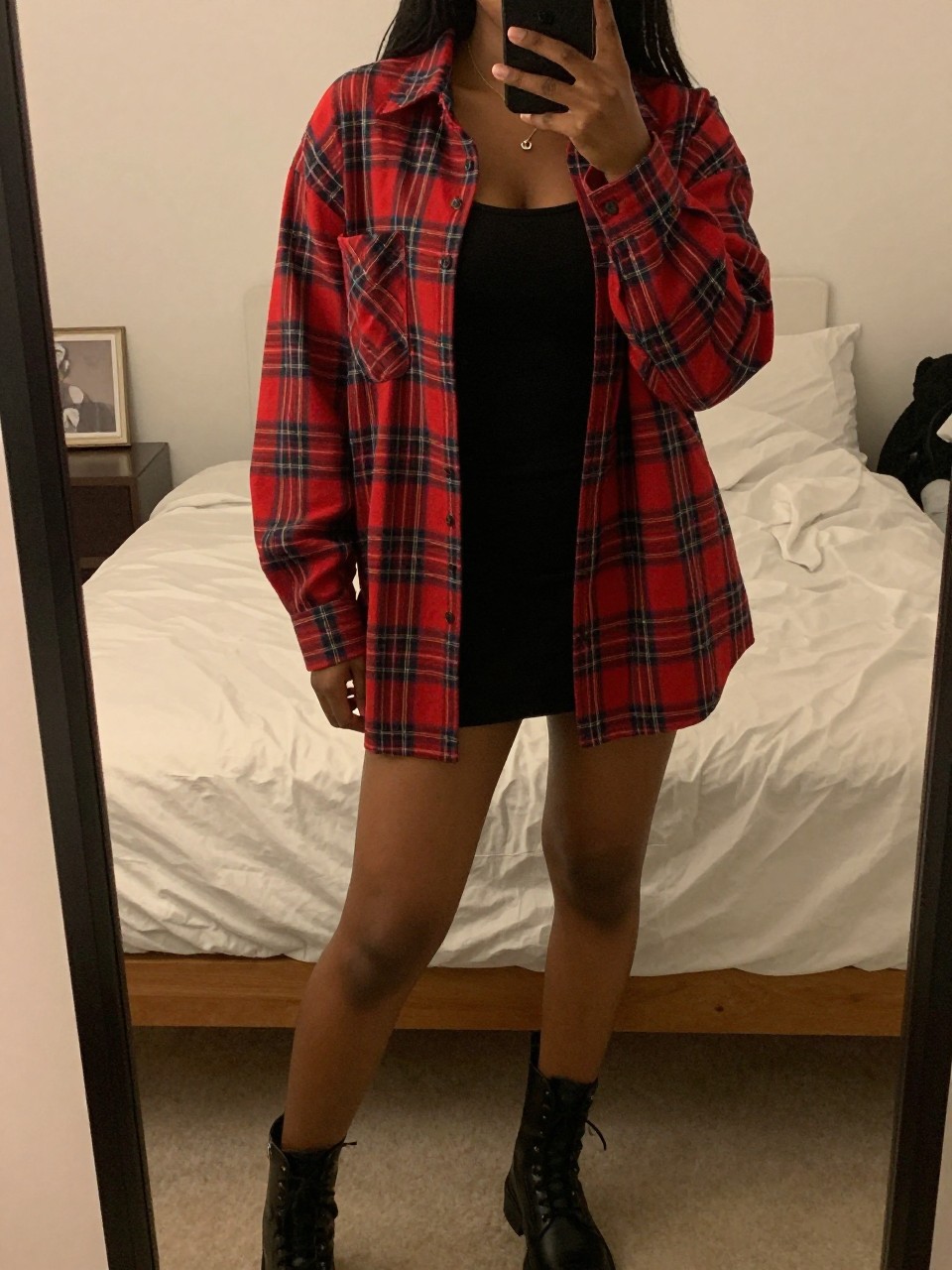 iPhone mirror selfie of a black woman wearing an oversized red plaid shirt open over a black tank top with a black mini skirt and black combat boots, one hand in her pocket, cropped at NECK so face doesn't show, in a feminine-themed bedroom, warm moody lighting, iPhone photo quality.