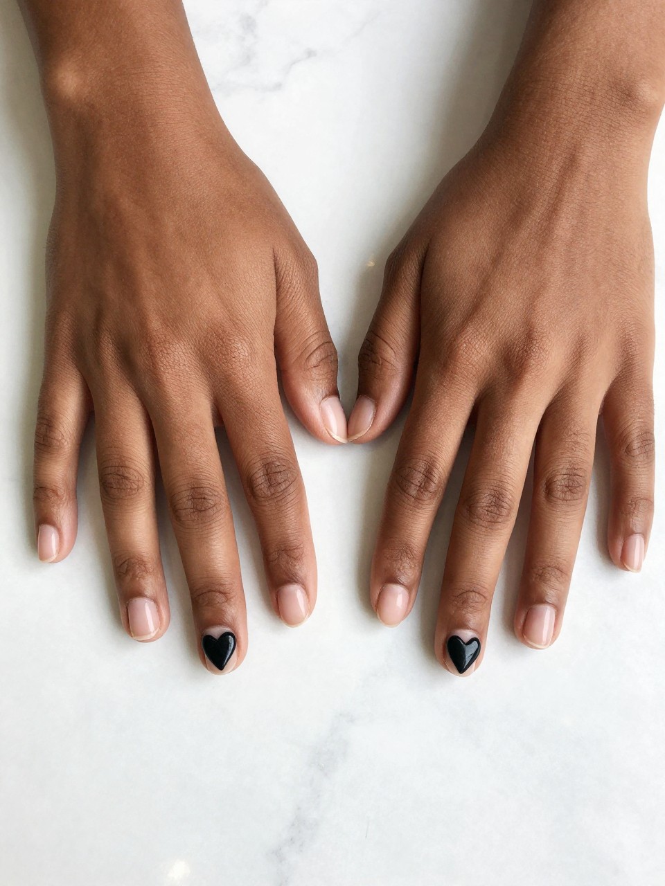 Top-down view of black feminine hands with natural, short nails on a white marble slab. A solid black heart sits each nails design. The look is clean, artistic, and slightly unsettling.