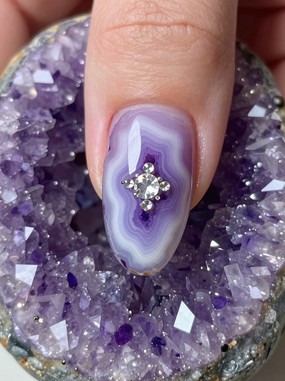 Macro, top-down view of a nail designed like a geode slice. Swirls of amethyst purple and white create a crystalline pattern. Small, clear rhinestones are clustered in the center. The hands rest on a rough amethyst stone, with light catching the crystals.