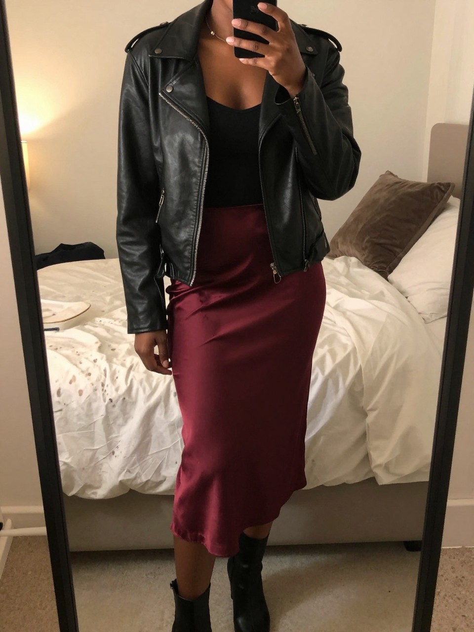 iPhone mirror selfie of a black woman wearing a black leather jacket over a black tank top with a burgundy satin slip skirt and black ankle boots, one hand in her jacket pocket, cropped at NECK so face doesn't show, in a feminine-themed bedroom, evening golden hour, iPhone photo quality.