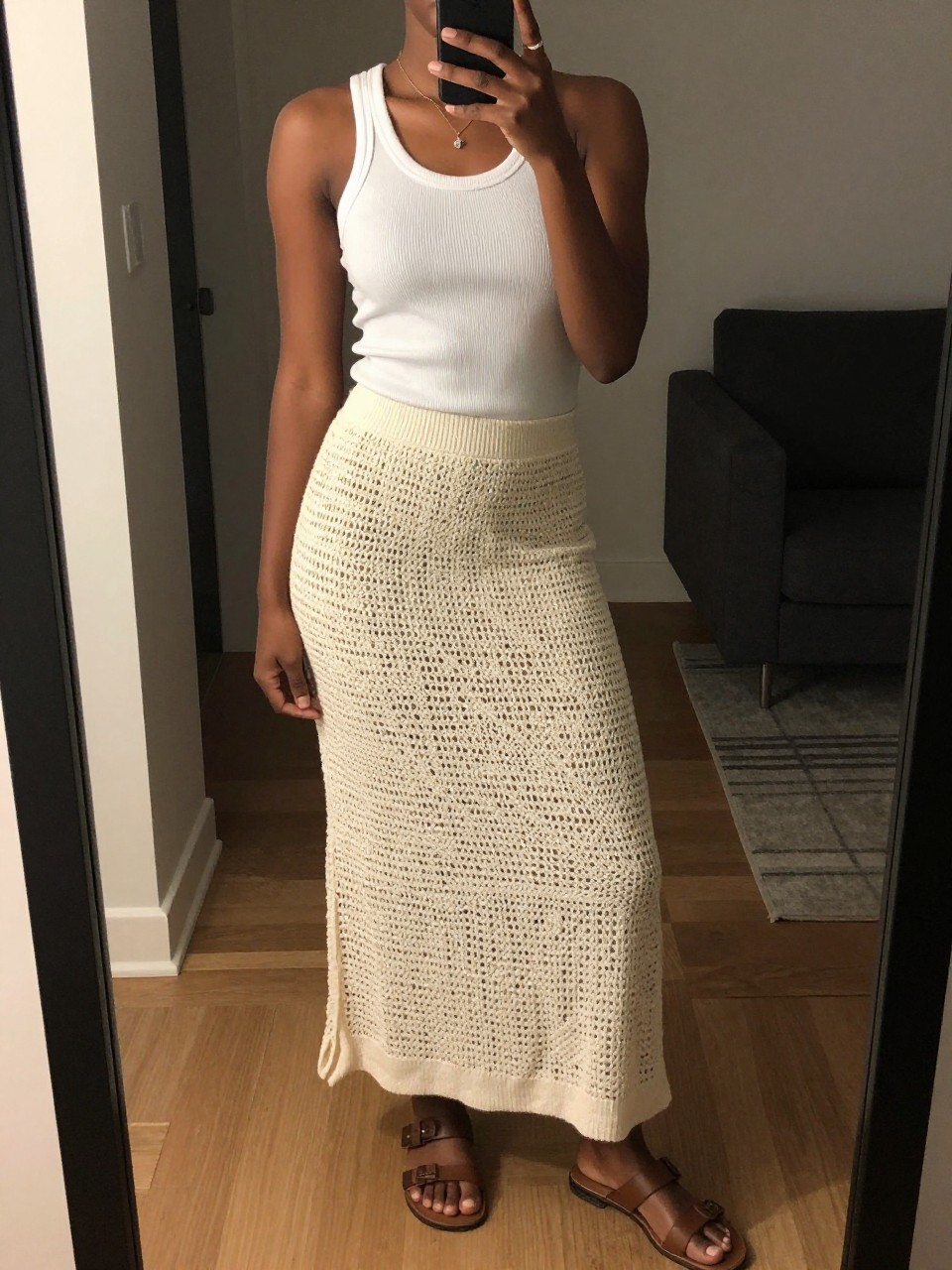 iPhone mirror selfie of a black woman wearing a simple white ribbed tank top with a cream crochet maxi skirt and flat brown leather sandals, one hand holding her phone at her side, cropped at NECK so face doesn't show, in a city apartment, warm moody lighting, iPhone photo quality.