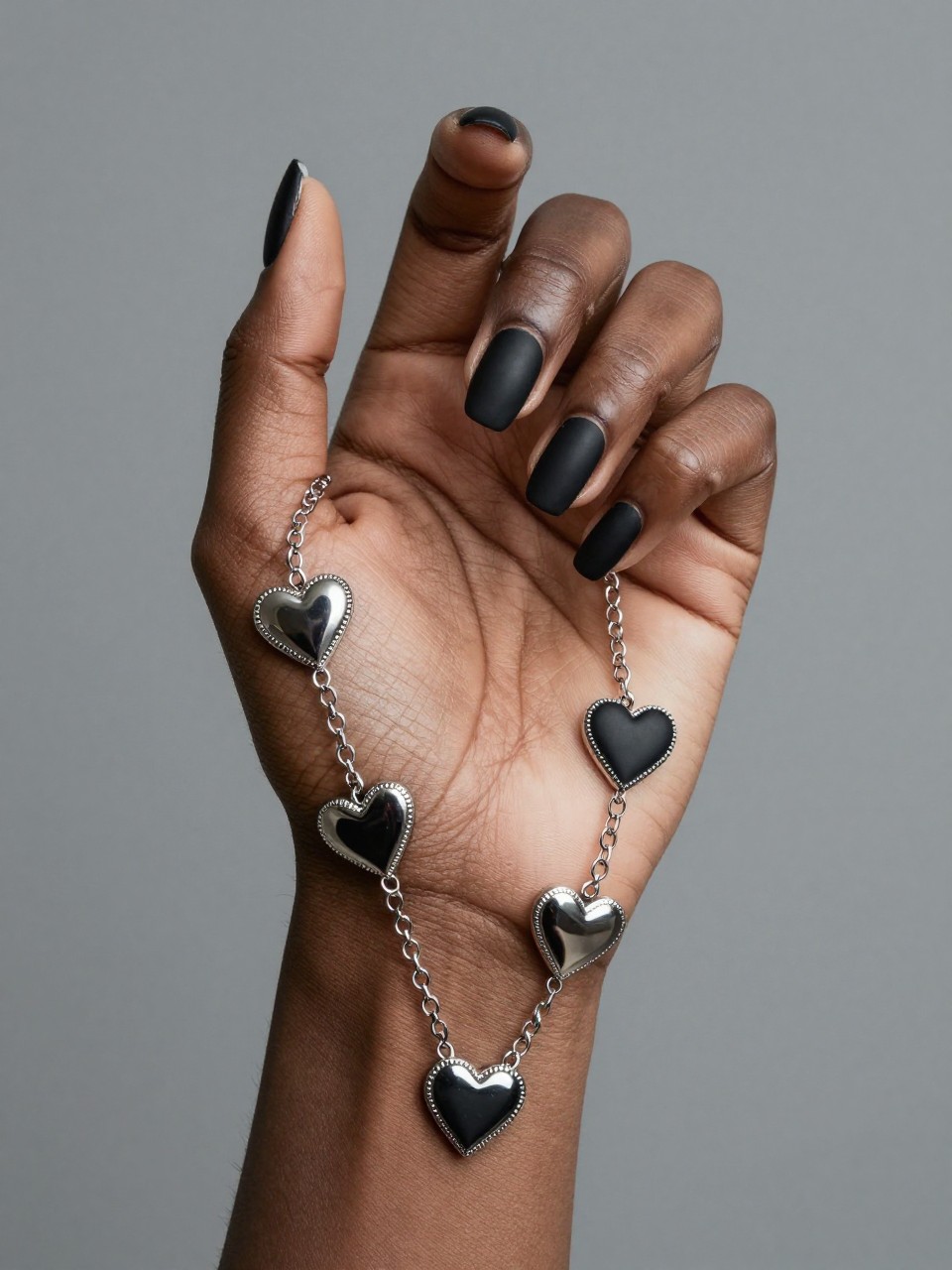 Creative shot of a black feminine hand with fingers slightly curled to show the flow of design. Chrome hearts and connecting chains are painted across all nails on one hand, creating a continuous "sleeve" effect on a matte black base. The look is cohesive and daring.