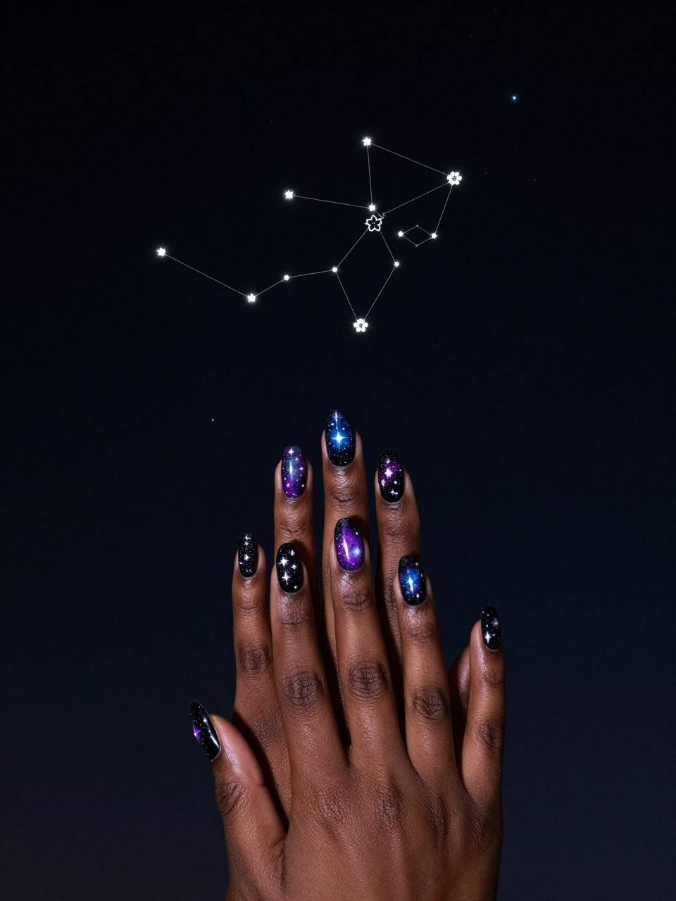 Bird's-eye view of black feminine hands with long nails against a dark backdrop, as if viewing the night sky. The nails are a galaxy of black, purple, and blue with silver glitter stars. Subtle white lines connect stars into tiny floral constellations. The mood is magical and vast.