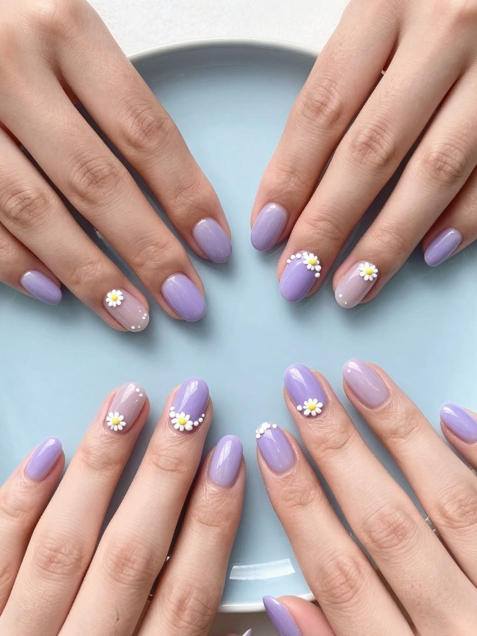 Overhead shot of hands with neat, oval nails on a light blue ceramic plate. The nails have a glossy periwinkle base. A single line of connected tiny white daisies wraps around each nail like a delicate chain. The composition is crisp and cute.