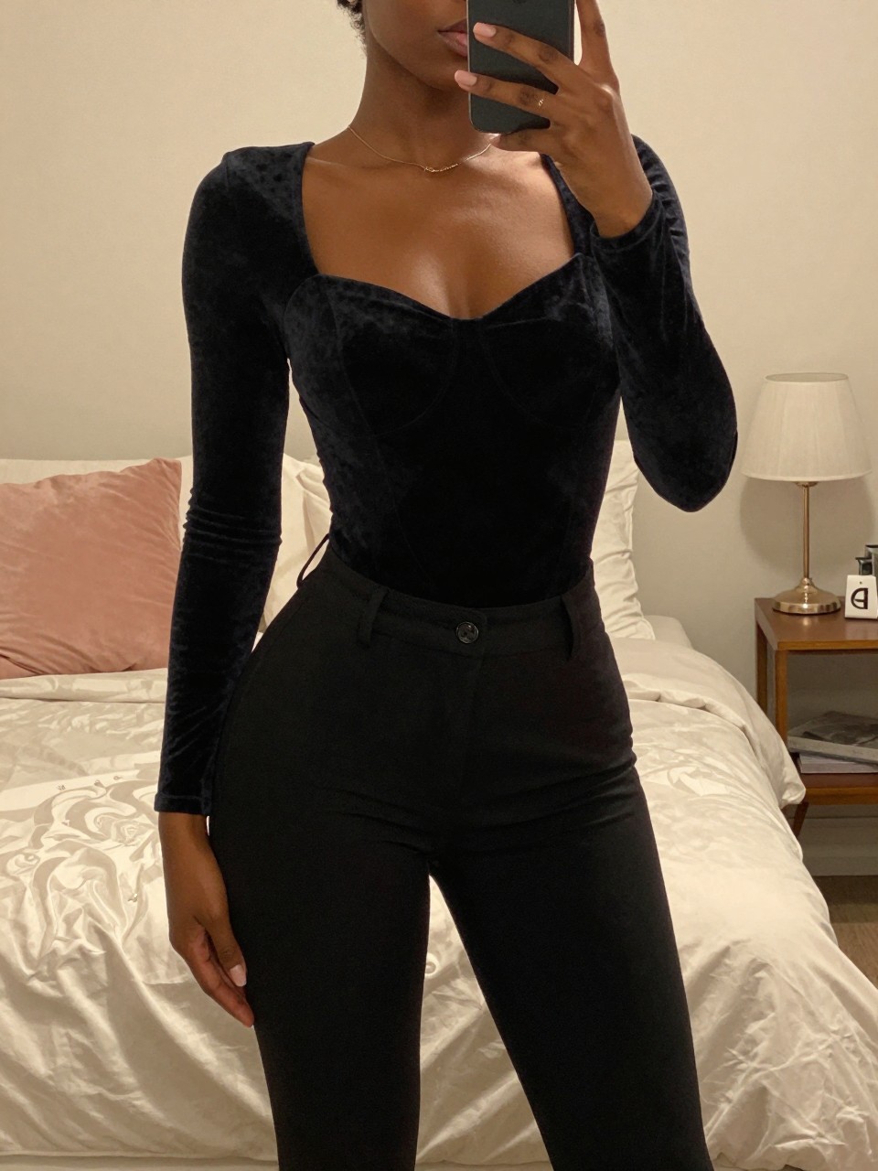 iPhone mirror selfie of a black woman in a velvet bodysuit and black trousers, posed with slight turn, cropped at NECK so face doesn't show, in a feminine-themed bedroom with warm moody lighting, iPhone photo quality.