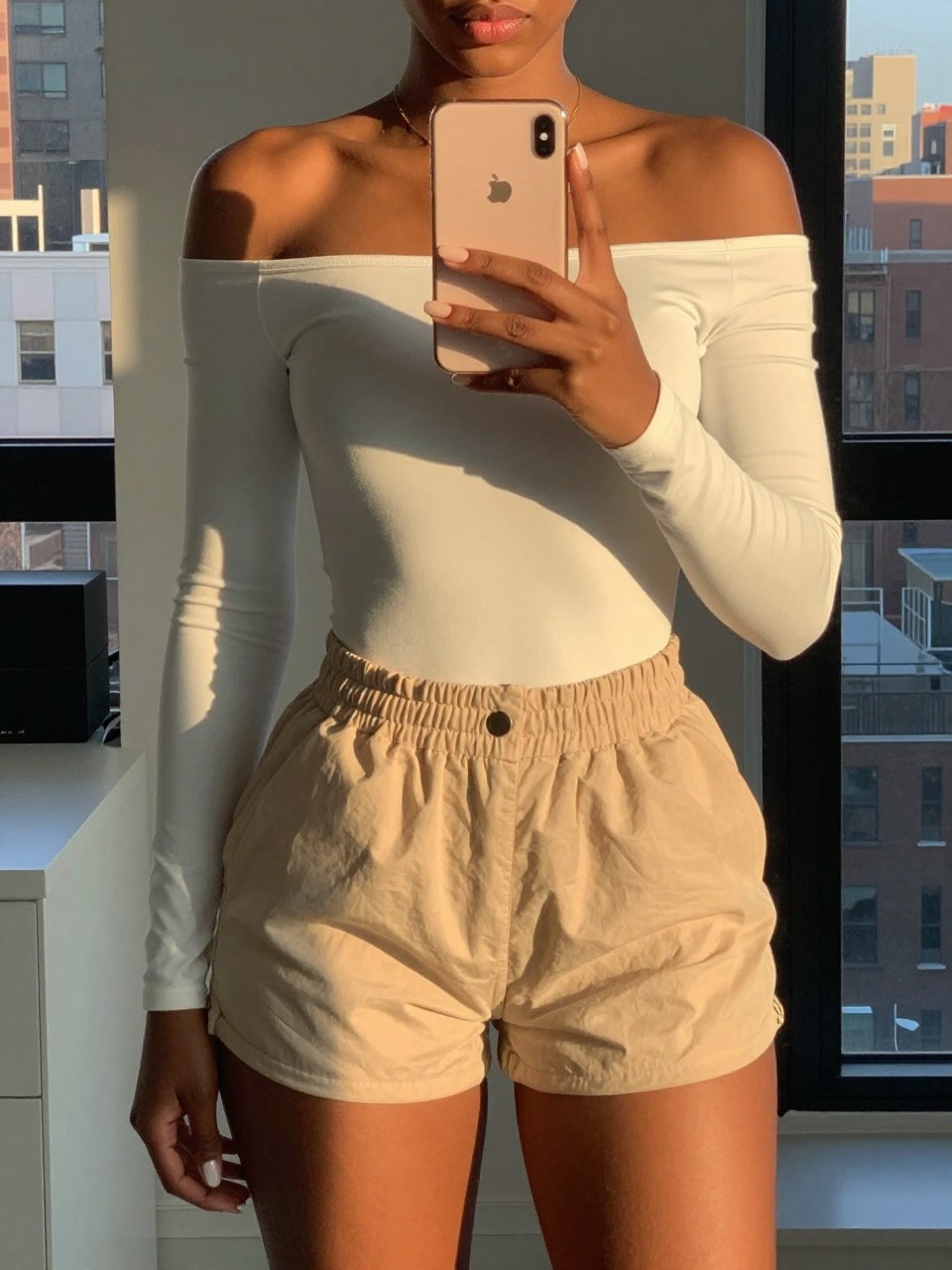 iPhone mirror selfie of a black woman in an off-shoulder bodysuit and paperbag shorts, holding phone at chest level, cropped at NECK so face doesn't show, in a city apartment during evening golden hour, iPhone photo quality.