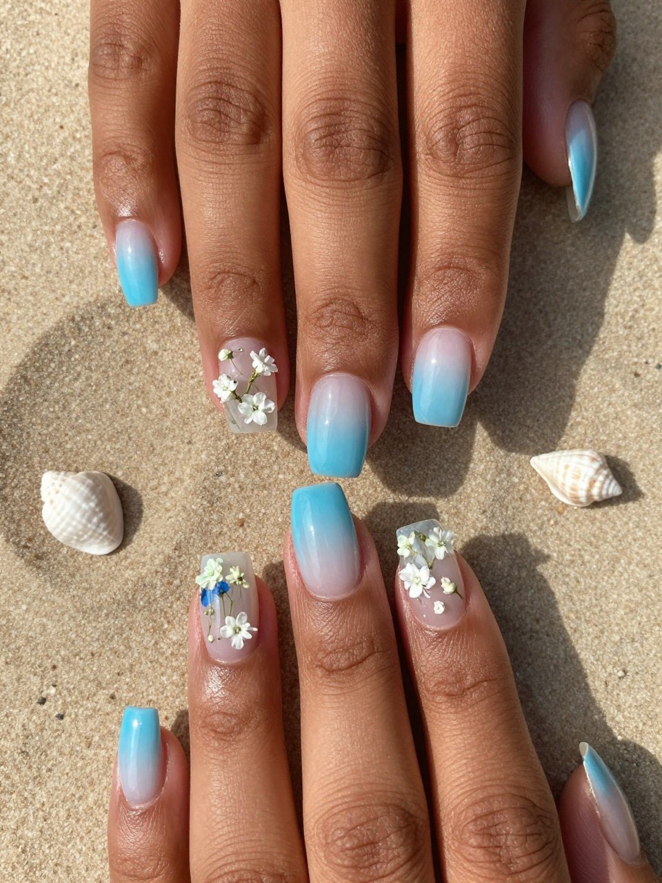 Macro, top-down view of black feminine hands with clear, glossy nails over a sand background. The nails have a sheer ocean blue tint. The ring fingers have tiny white seashells and pressed baby’s breath flowers sealed under the gel, like treasures in resin.