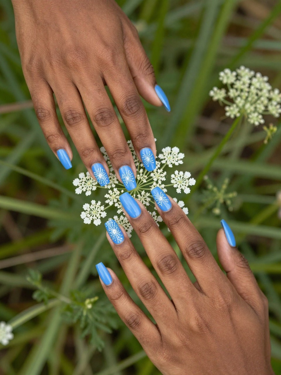Overhead view of black feminine hands with long nails gently touching a sprig of Queen Anne's Lace. The nails are a bright cornflower blue. Each nail features a delicate, dot-created white lace flower pattern. The ambiance is bright and meadow-like.