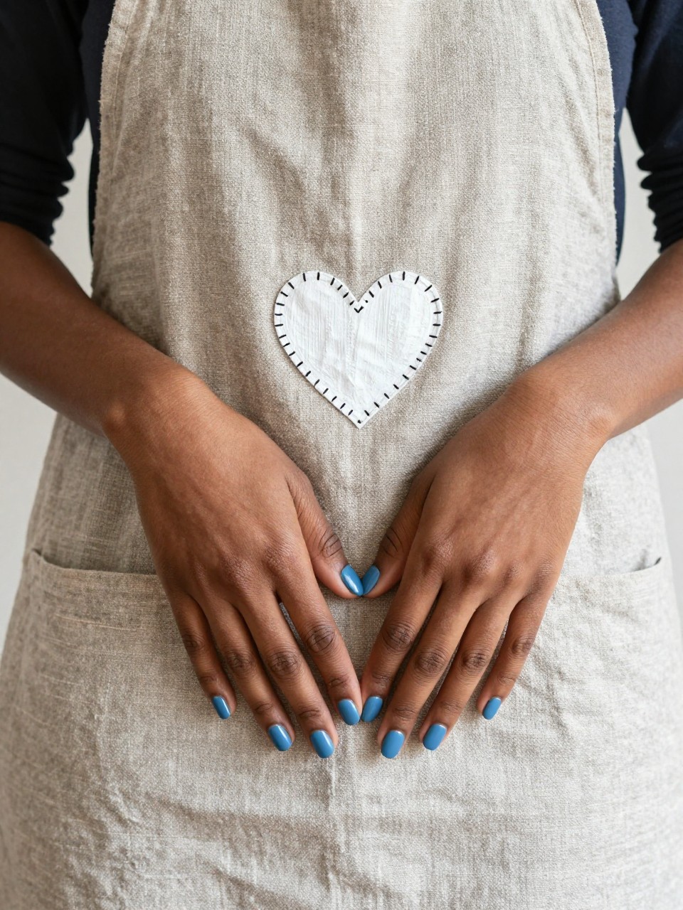 Bird's-eye view of black feminine hands resting on a rustic linen apron. The nails are a matte chambray blue. A white heart is painted with small black "stitch" marks around its border. The vibe is handmade and cozy.