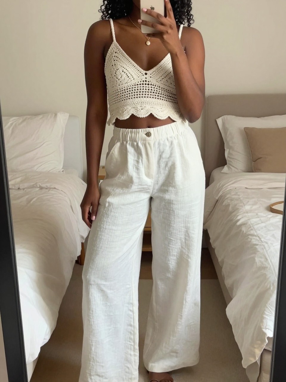iPhone mirror selfie of a bklack woman wearing white linen wide-leg pants and an ecru crochet top, standing casually with one hand holding her phone, cropped at NECK so face doesn't show, in a bright feminine-themed bedroom, warm moody lighting, iPhone photo quality.