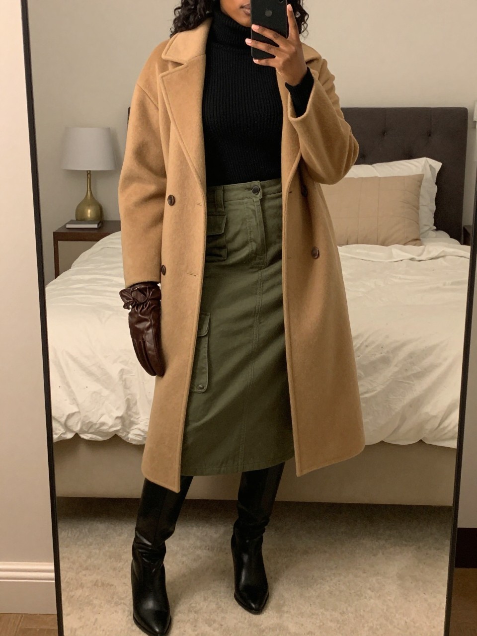iPhone mirror selfie of a black woman wearing a black fine-knit turtleneck tucked into an olive green cargo midi skirt with a long camel wool coat over top and black knee-high boots, holding leather gloves, standing in an elegant winter pose, cropped at NECK so face doesn't show, in a feminine-themed bedroom, warm moody lighting, iPhone photo quality.