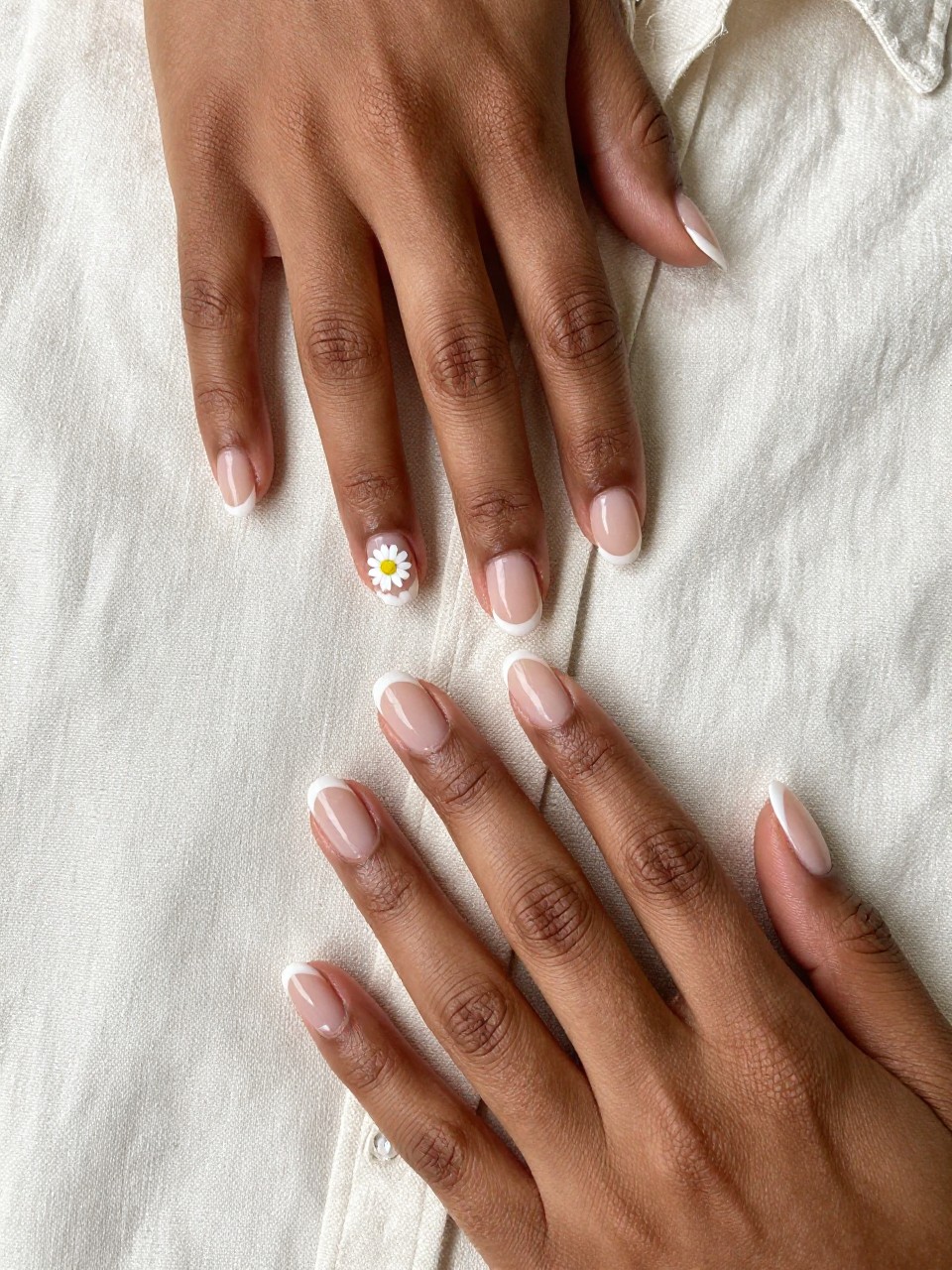 Close-up, top-down view of black woman hands with neat, natural-length nails on a beige linen shirt. The nails have a sheer nude base. One tiny daisy is painted near the base of each nail. The look is pristine, understated, and chic.