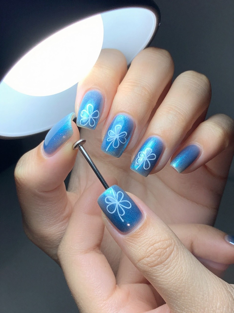 Overhead shot of hands with coffin nails under a bright light, showing off a holographic blue polish shift. Each nail features a simple, white outline drawing of a flower. The look is modern, magical, and eye-catching.
