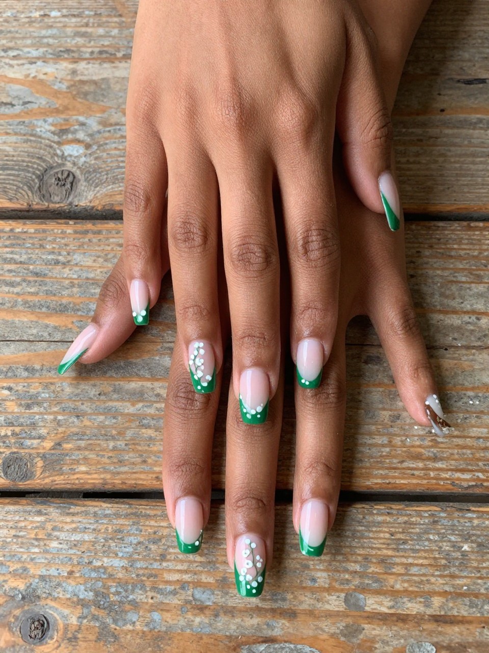 Top-down view of black feminine hands with long nails on a rustic wooden table. The French tip is a wavy, uneven green line. Small white dot flowers are painted along the green wave, making the tip itself look like a flowering vine.