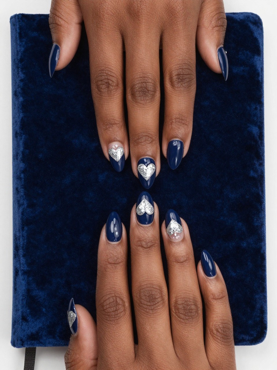 Bird's-eye view of black feminine hands with almond nails resting on a dark blue velvet journal. The nails are a glossy navy blue. Small, glittering silver hearts are placed on each nail. The lighting is soft, making the glitter hearts twinkle against the deep base.