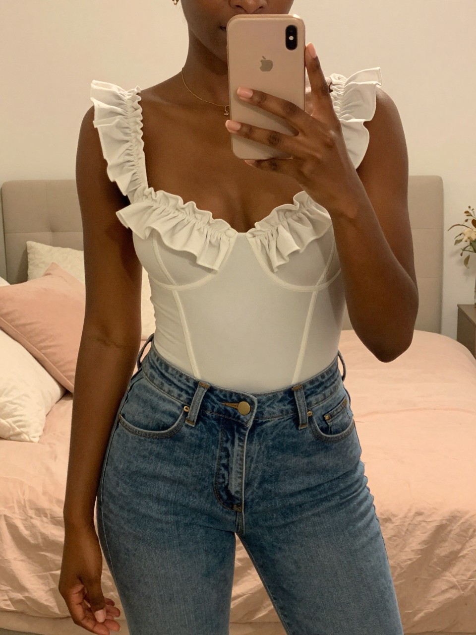 iPhone mirror selfie of a black woman in a ruffled bodysuit and denim jeans, phone held naturally, cropped at NECK so face doesn't show, in a feminine-themed bedroom with warm moody lighting, iPhone photo quality.