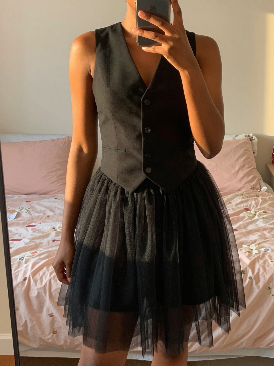 iPhone mirror selfie of a black woman wearing a black vest with a tulle skirt, standing with her phone hand at her side, cropped at NECK so face doesn't show, in a feminine-themed bedroom, evening golden hour light, iPhone photo quality.
