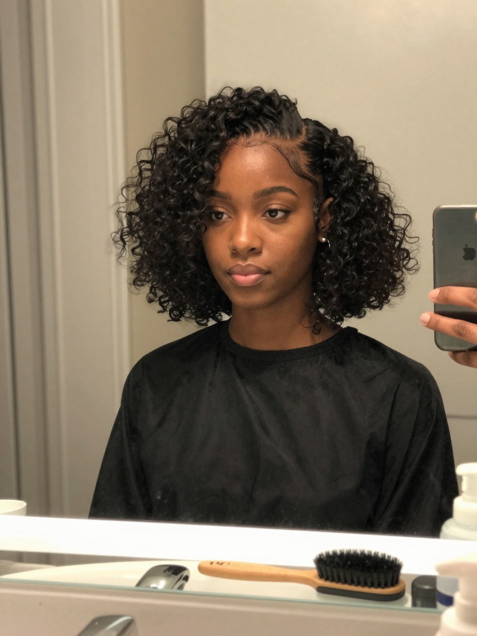 Photo of a black woman wearing a curly bob with a deep side part, front-facing view showing the volume on one side, setting is a bathroom vanity mirror with soft indoor lighting, containing an edge brush on the counter, iPhone photo quality.