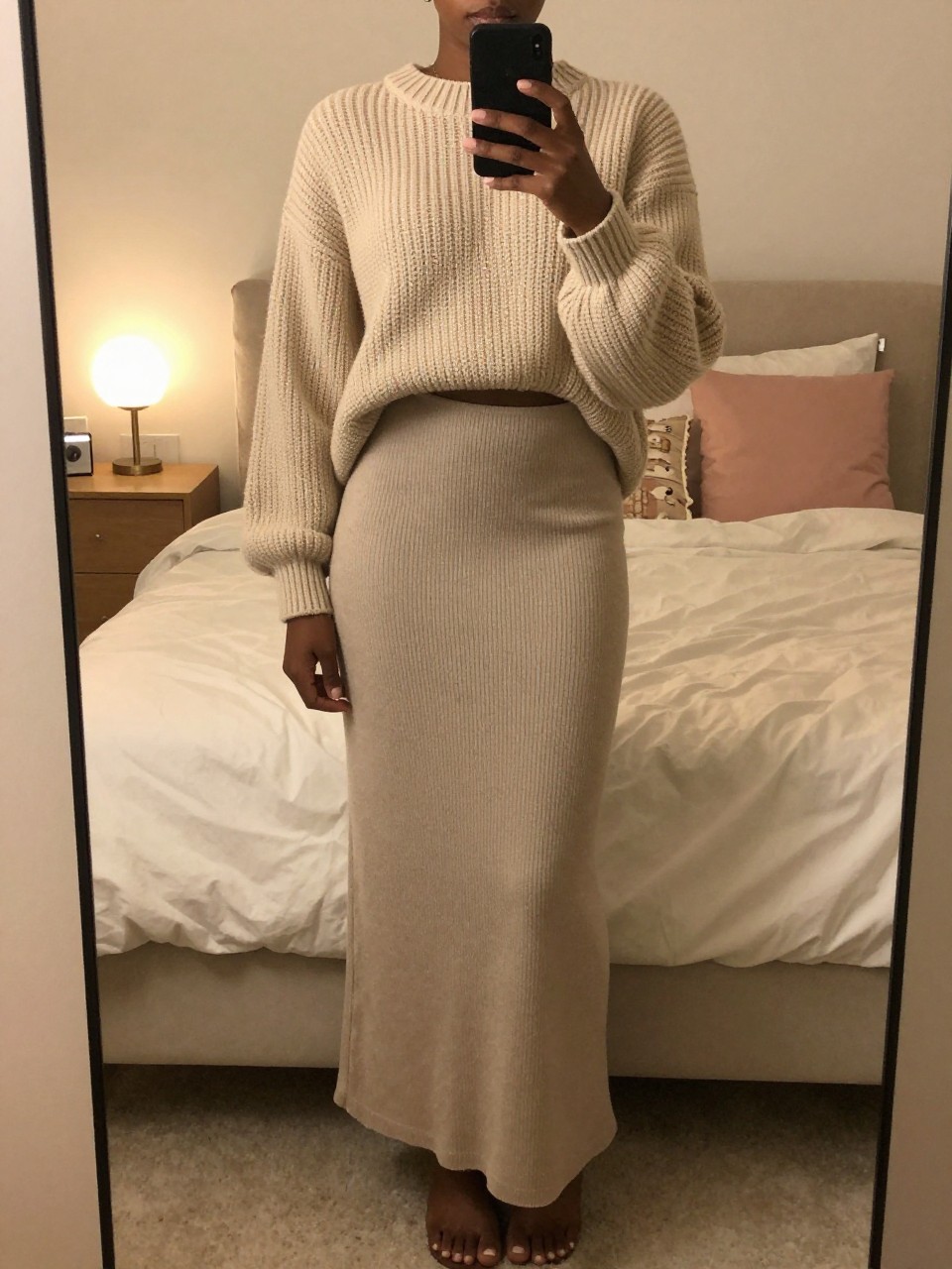 iPhone mirror selfie of a black woman in a maxi skirt and chunky sweater with Timbs, holding phone low to capture length, cropped at NECK so face doesn't show, in a feminine-themed bedroom with warm moody lighting, iPhone photo quality.