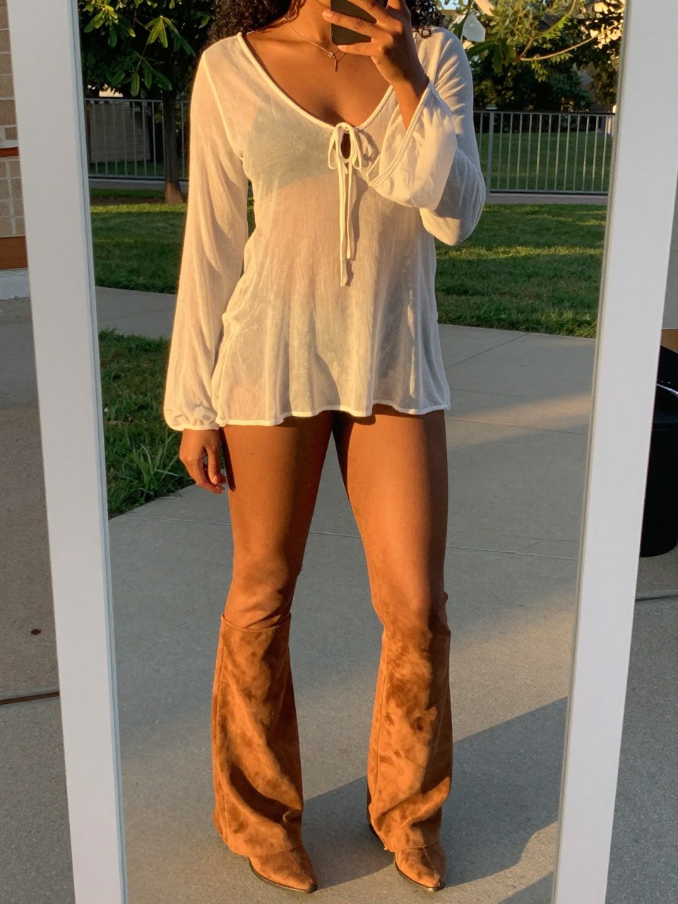 mirror selfie of a black woman in a sheer boho top and suede flares, angled slightly, cropped at NECK so face doesn't show, in a sunny outdoor setting during evening golden hour, iPhone photo quality.