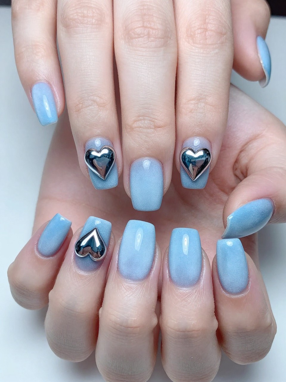 Close-up, top-down view of hands under cool, bright lighting. The nails are a frosty light blue. Perfect, mirror-like chrome hearts are centered on each nail, reflecting the surroundings. The look is sleek, modern, and frosty.