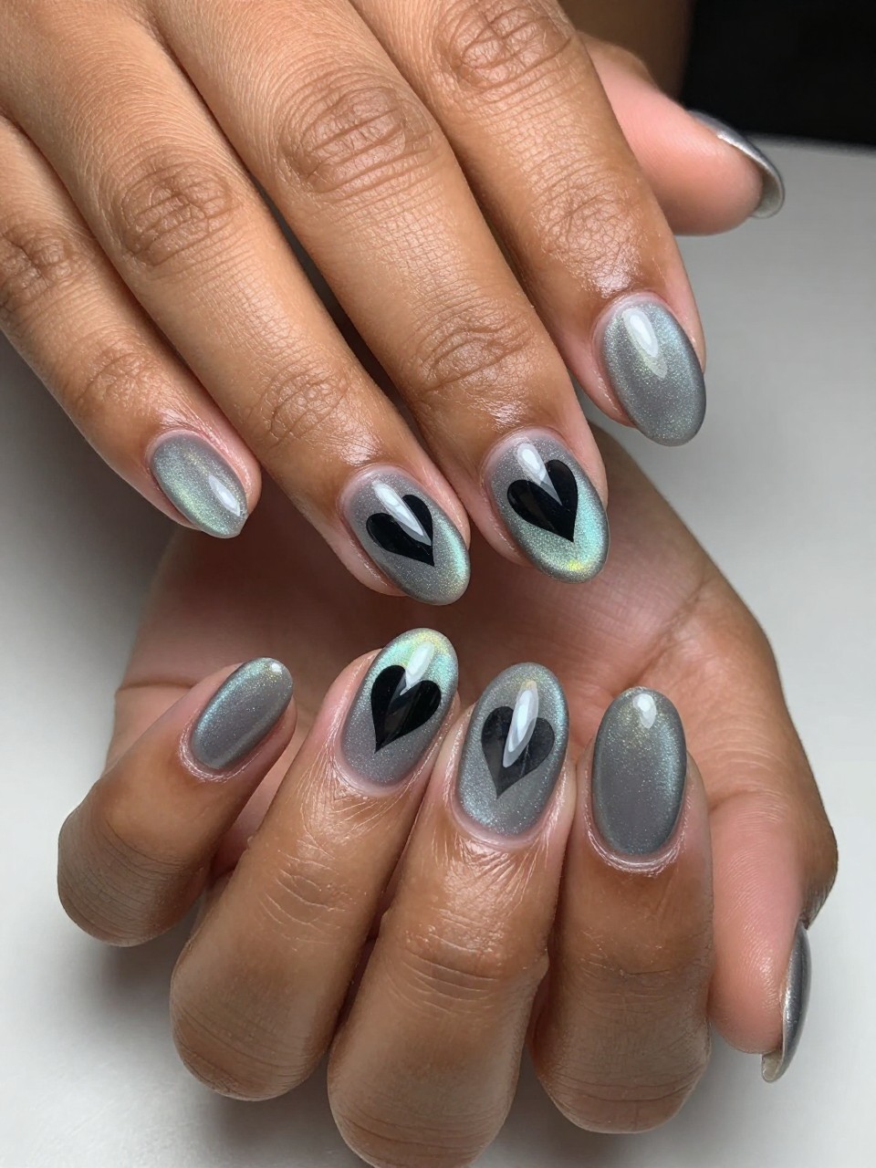Angled view of black feminine hands with oval nails under shifting light. The base is a smoky holographic grey cat eye. A solid black heart silhouette appears on each nail, unaffected by the surrounding shimmer. The effect is mesmerizing and modern.