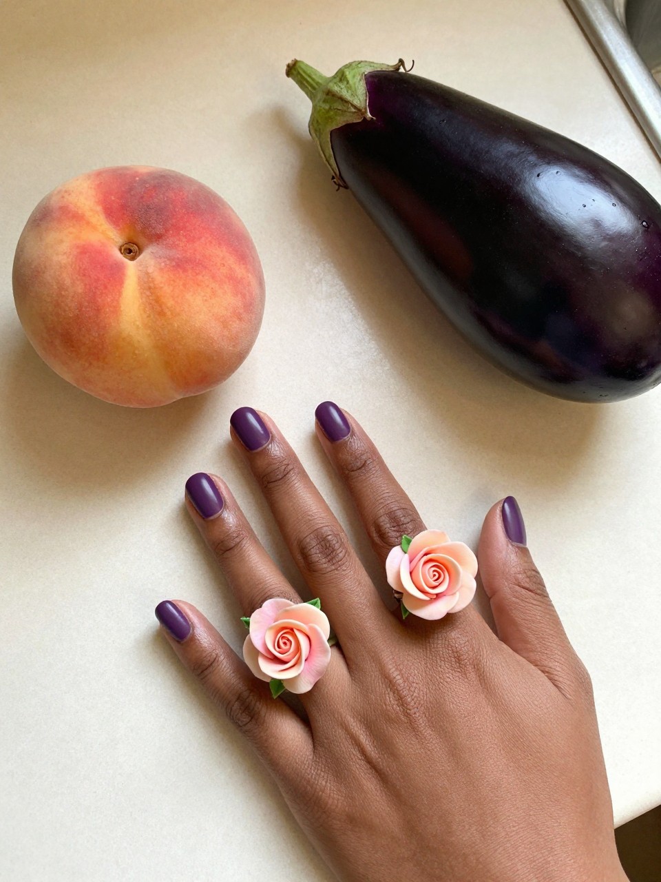 Overhead view of black feminine hands with rounded nails next to a peach and an eggplant on a kitchen counter. The nails are a matte eggplant purple. Each ring finger design features a single, meticulously painted peach rose. The lighting is warm and natural.