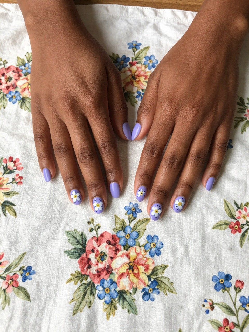 Bird's-eye view of black feminine hands with short nails resting on a vintage floral handkerchief. The nails are a matte lavender. Tiny blue and white forget-me-nots with yellow centers are painted, surrounded by small black “stitch” marks. The vibe is crafty and sentimental.