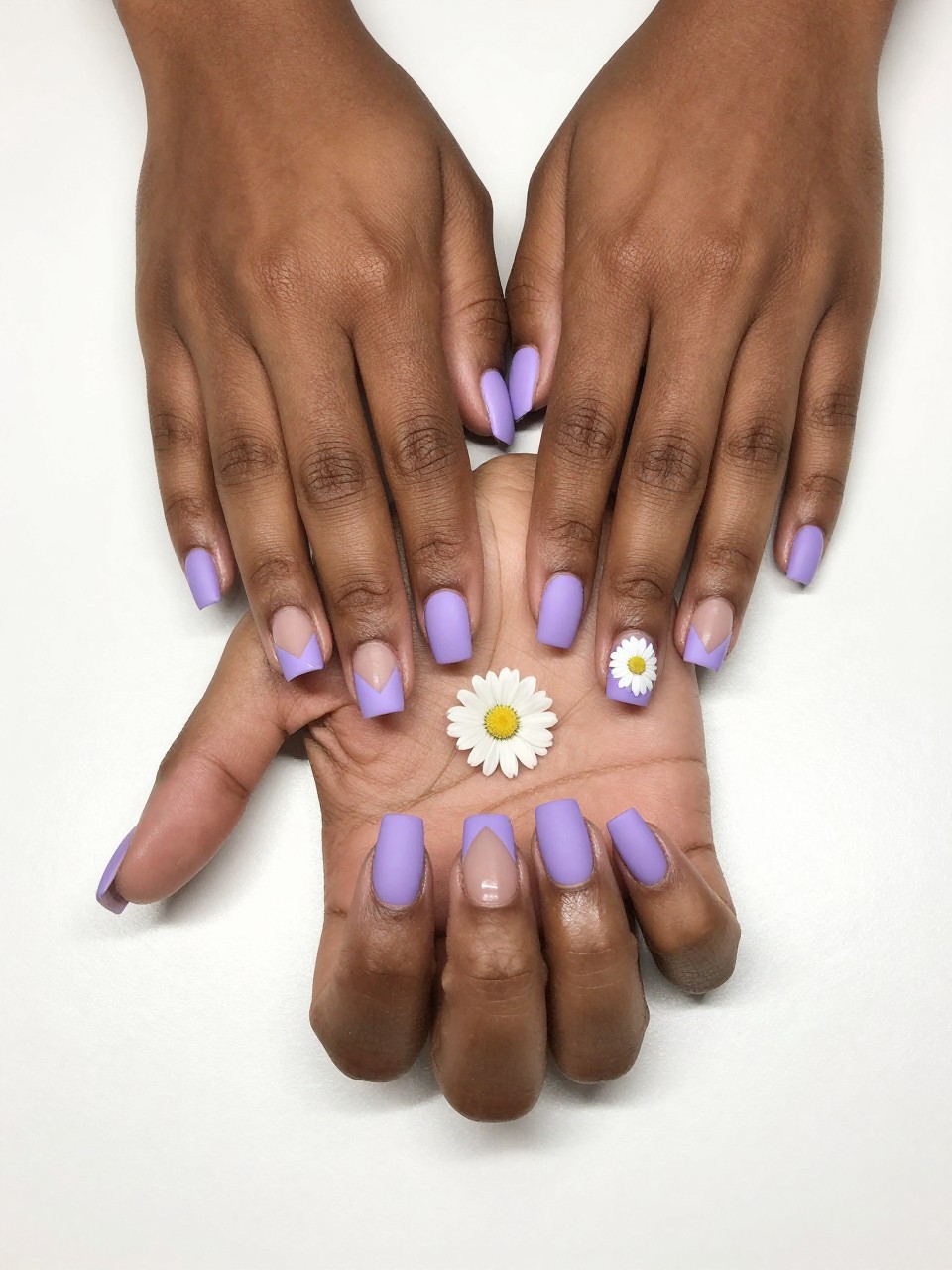 Top-down view of black feminine hands with short, squared nails on a white desk. The nails are mostly matte pastel purple, but each has a sharp triangular cut-out of bare nail at the side. A single, clean white daisy is painted inside that triangle. The look is architectural.