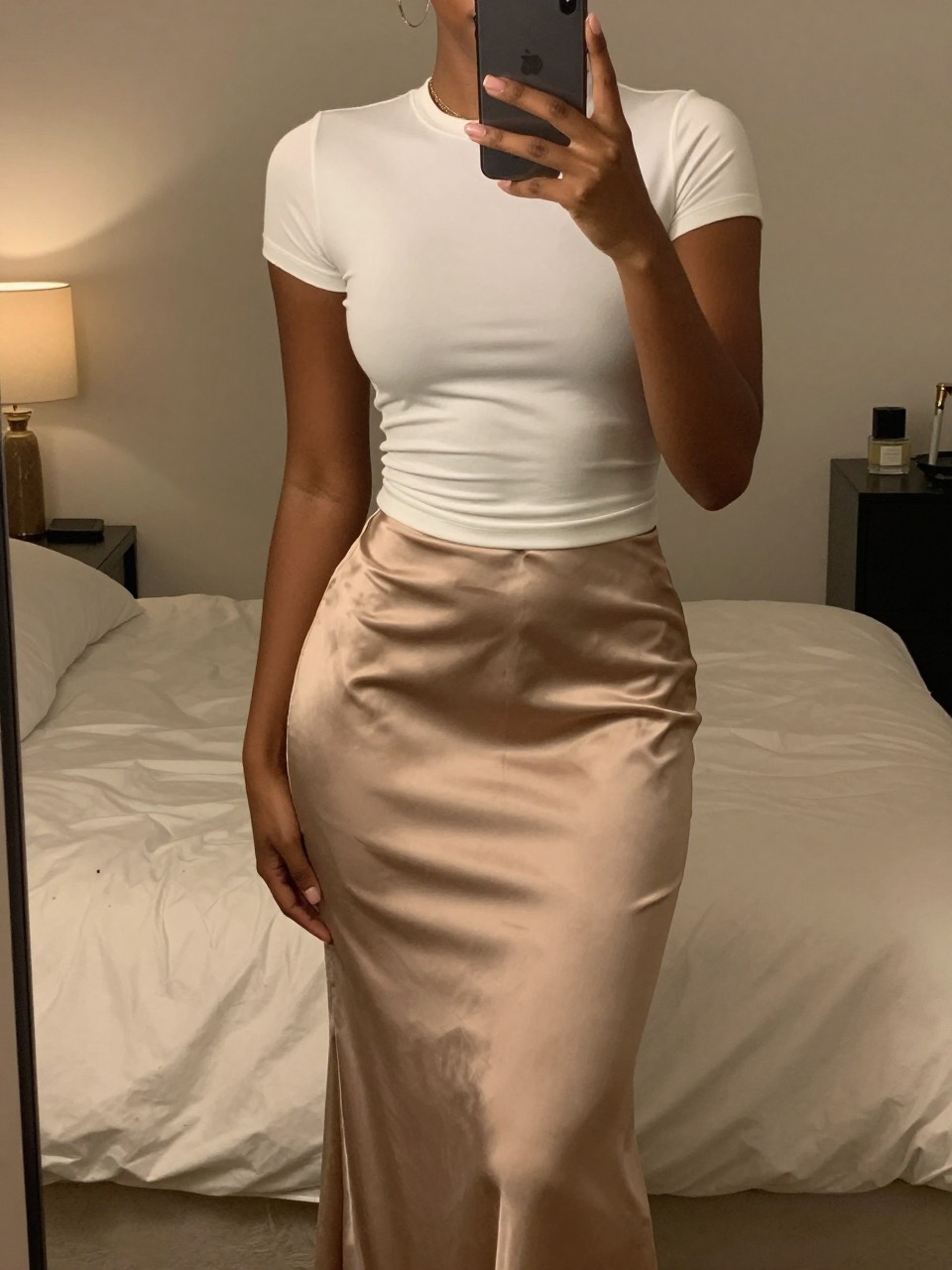 iPhone mirror selfie of a black woman in a satin maxi skirt and fitted tee, phone at natural height, cropped at NECK so face doesn't show, in a feminine-themed bedroom with warm moody lighting, iPhone photo quality.