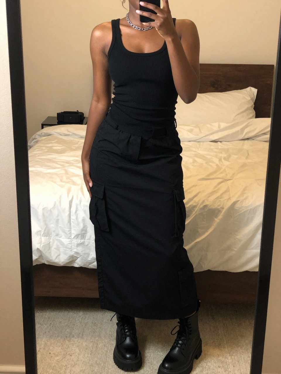 iPhone mirror selfie of a black woman wearing a black ribbed tank top tucked into a black cargo maxi skirt with chunky black combat boots and a silver chain necklace, standing in an edgy pose with hands in pockets, cropped at NECK so face doesn't show, in a feminine-themed bedroom, warm moody lighting, iPhone photo quality.
