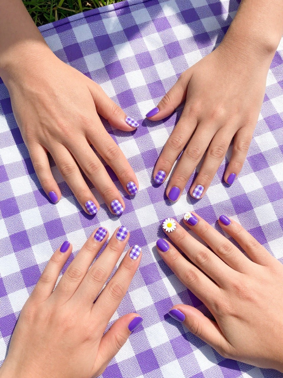 Top-down view of hands with short nails on a purple and white gingham picnic blanket. Some nails are purple and white checkered. The accent nails are solid purple with one bold white daisy. The setting is a sunny park.