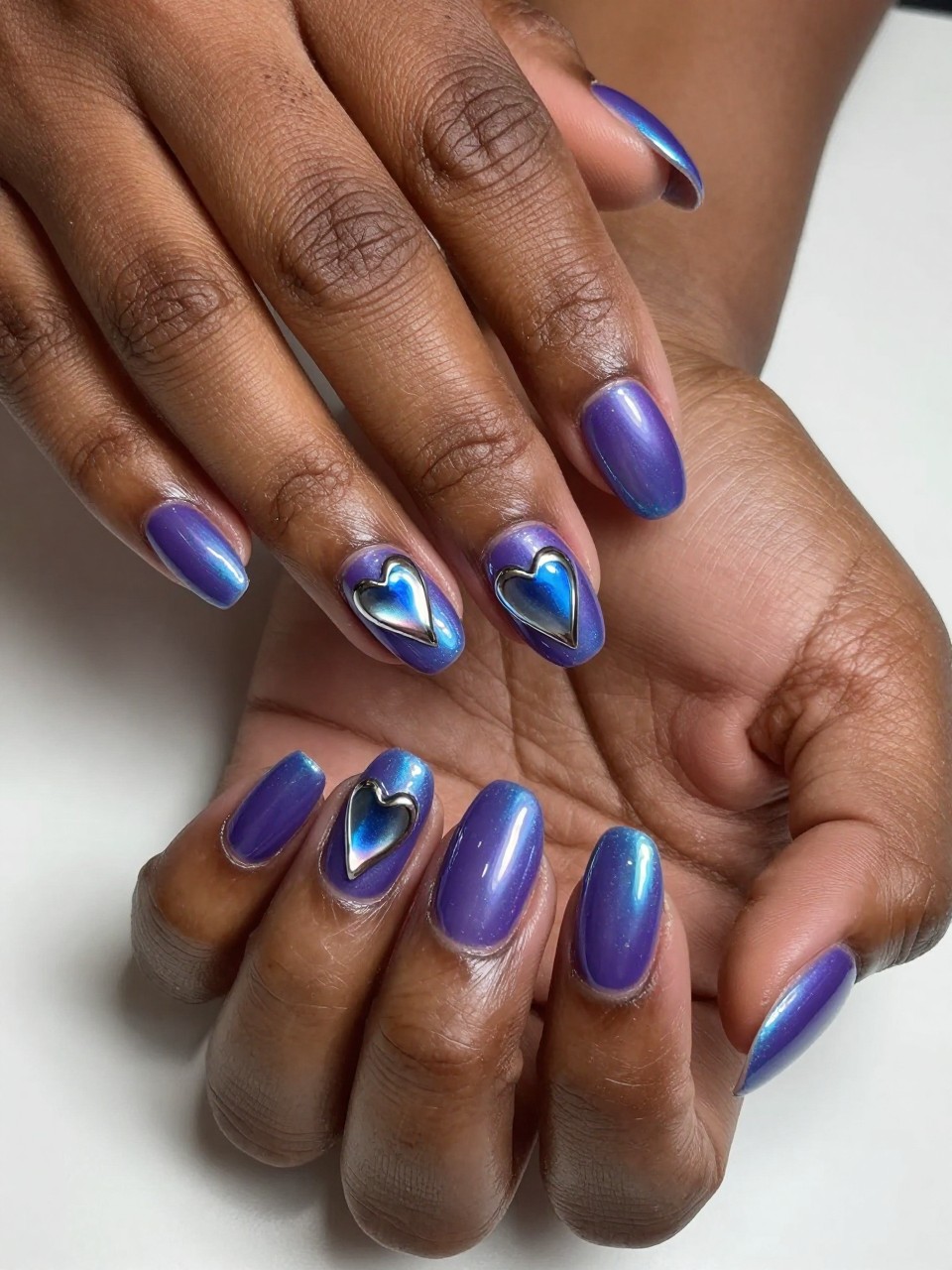 Angled overhead shot of black feminine hands under shifting light. The nails are a purple-to-blue duochrome. The chrome hearts, made from the same pigment, blend seamlessly, only visible through their reflective intensity. The effect is liquid metal.