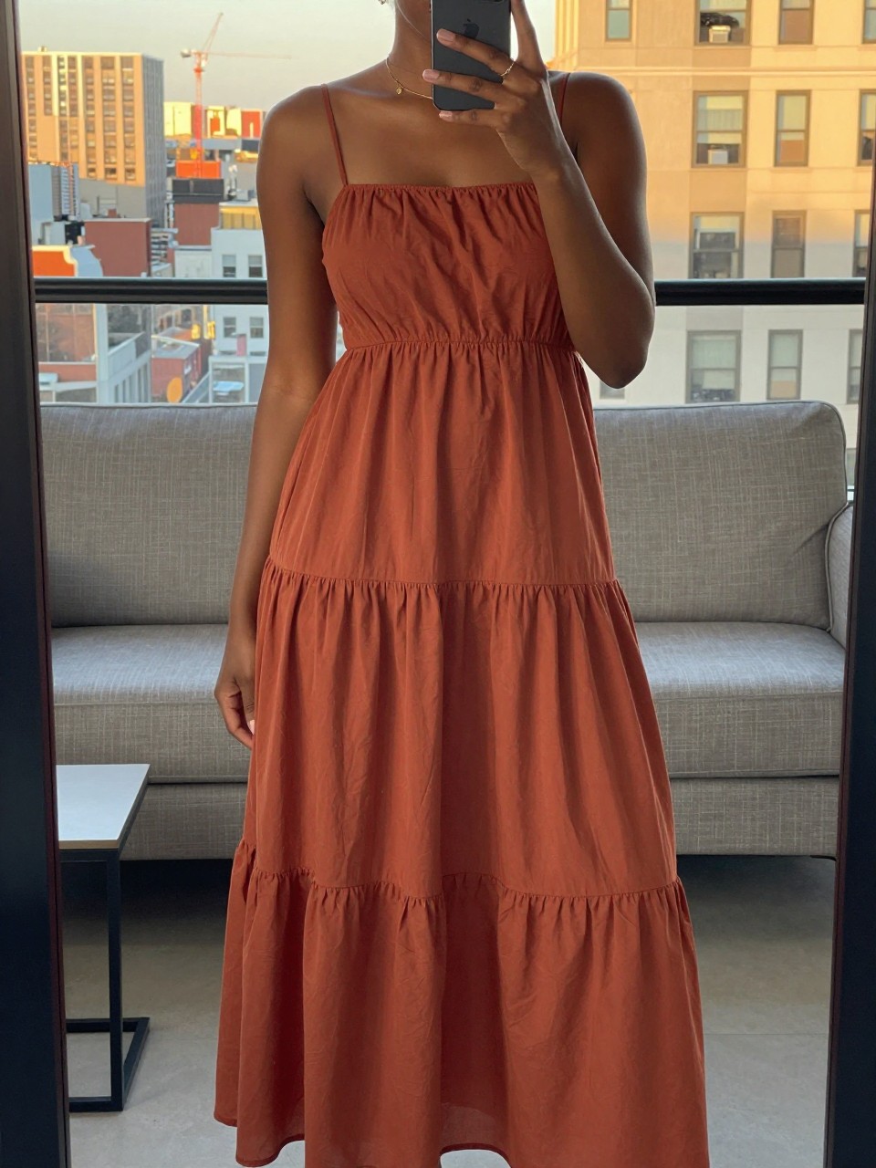 mirror selfie of a black woman in a terracotta tiered maxi dress, phone held naturally, cropped at NECK so face doesn't show, in a city apartment during evening golden hour, iPhone photo quality.