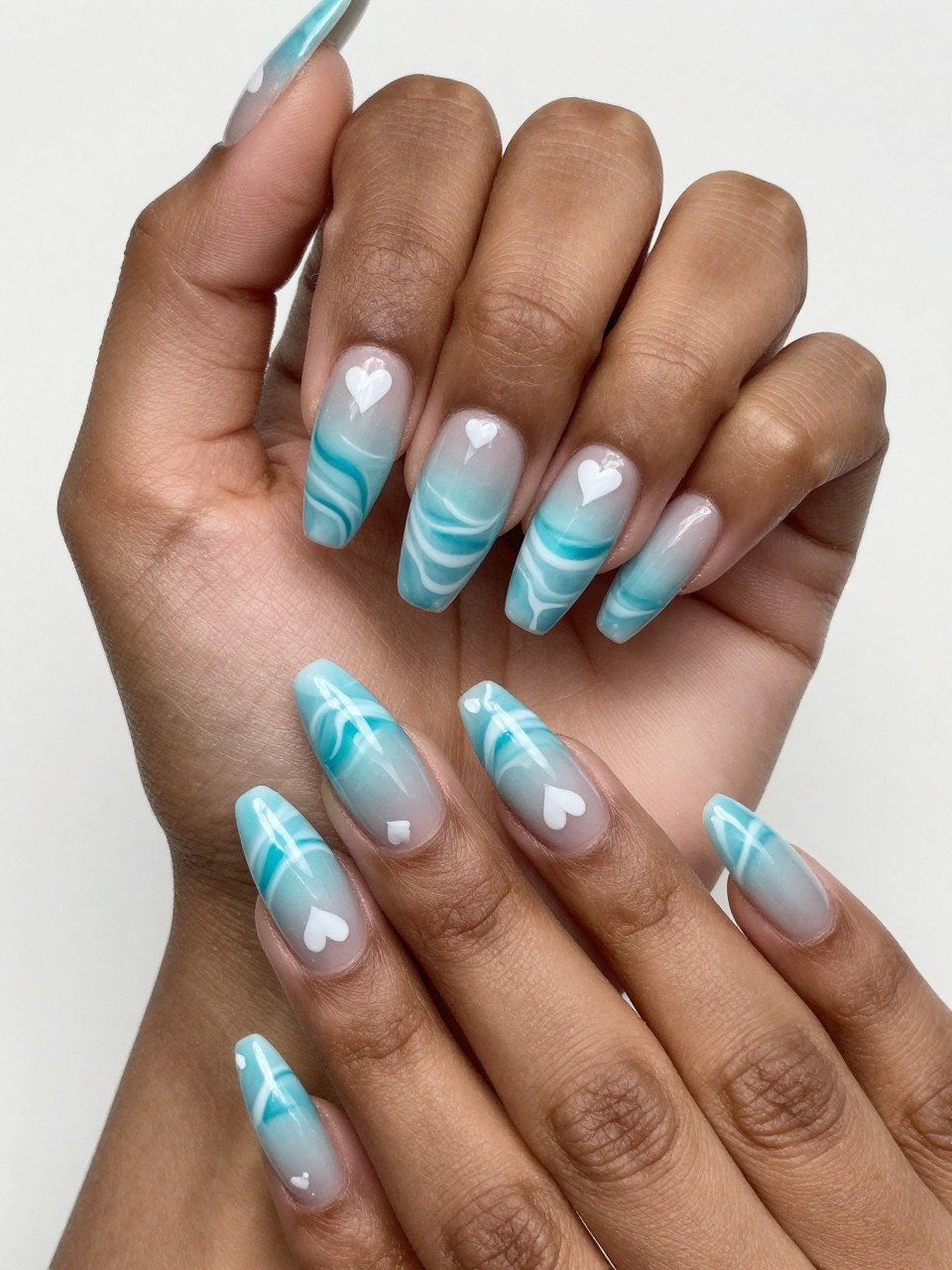 Angled view of black feminine hands with long nails. The nails design show a beautiful ocean wave ombré. Small white hearts are painted on it. The effect is serene and magical.