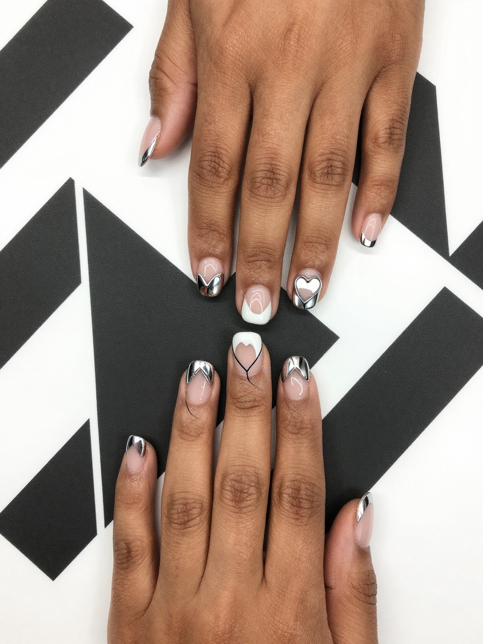 Top-down view of black feminine hands with a French manicure, but the tips are heart-shaped and made of mirror chrome. A hair-thin black outline traces the heart shape. Hands rest on a geometric black and white print, emphasizing the graphic quality.