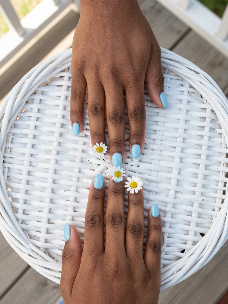 Overhead, top-down view of black feminine hands with short, oval nails resting on a white wicker basket. The nails are a soft, glossy baby blue. Three or four white daisies with yellow centers are dotted playfully on each nail. The setting is a bright, sunlit porch.